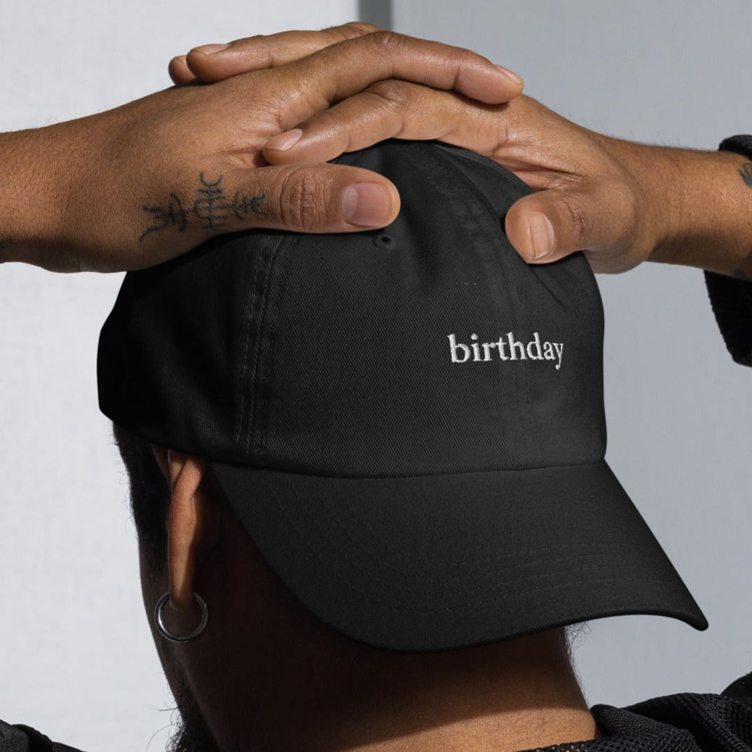 Person wearing a black cap with 'birthday' text, hands on head, against a neutral background