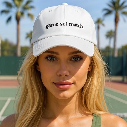Woman wearing a white cap with 'game set match' text on a tennis court with palm trees in the background